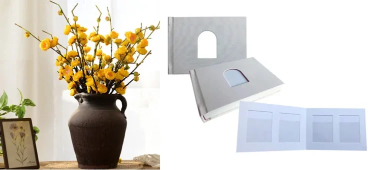 Grey photo album with a cutout window displayed next to vase with yellow flowers and framed art on a table.