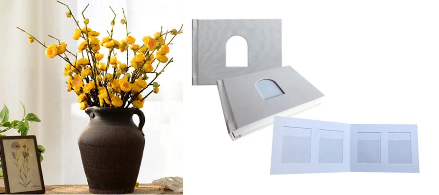 Grey photo album with a cutout window displayed next to vase with yellow flowers and framed art on a table.