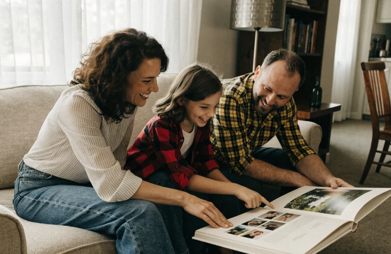 A family smiling as they look through a large, open coffee table album together on a sofa.